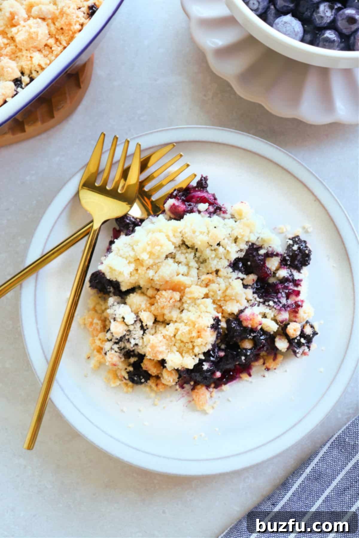 A serving of blueberry crumble on a small dessert plate, with two golden forks on the side.