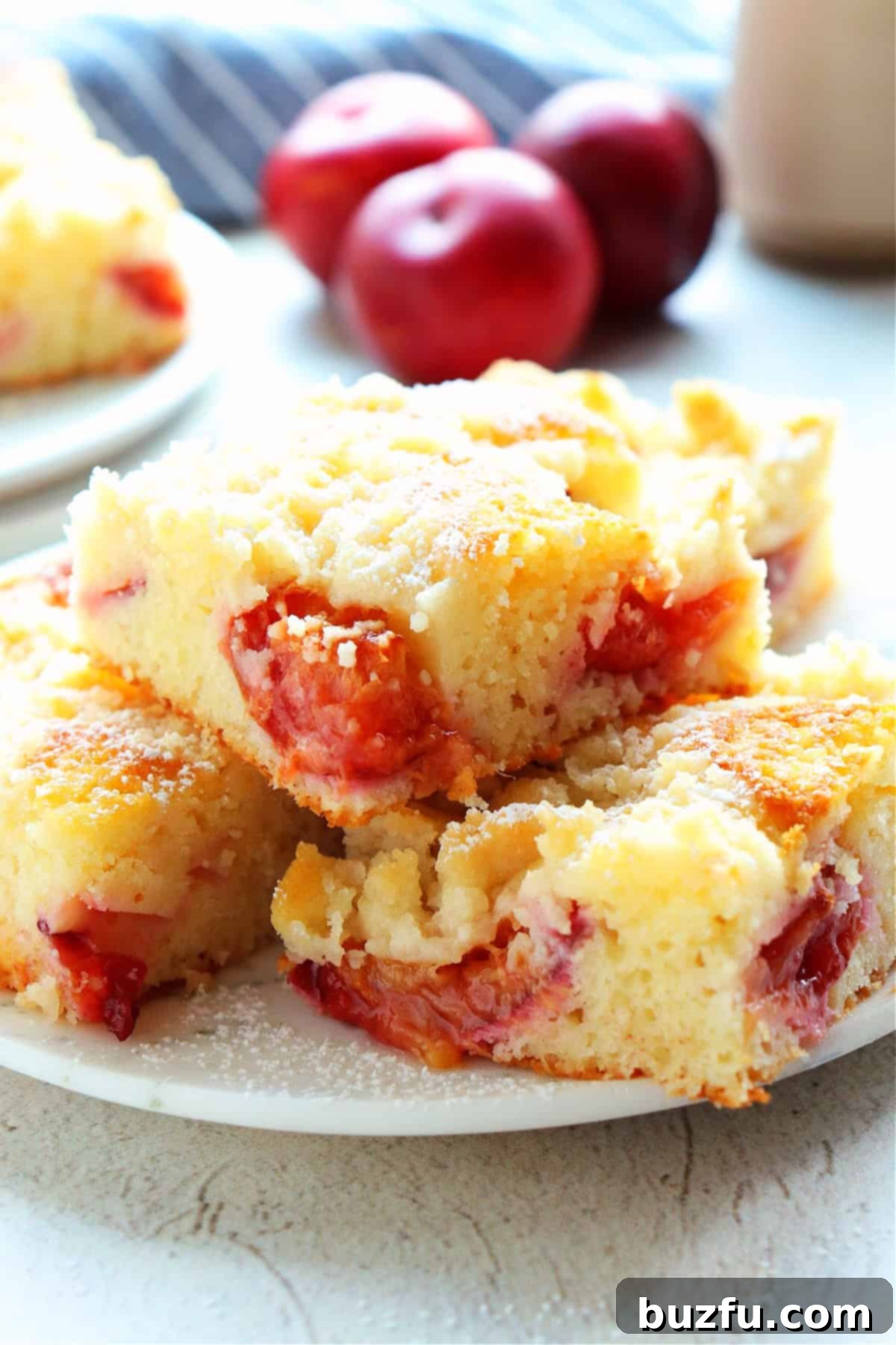 Close up photo of slices of coffee cake with plums on a white dessert plate, fresh plums in the back.