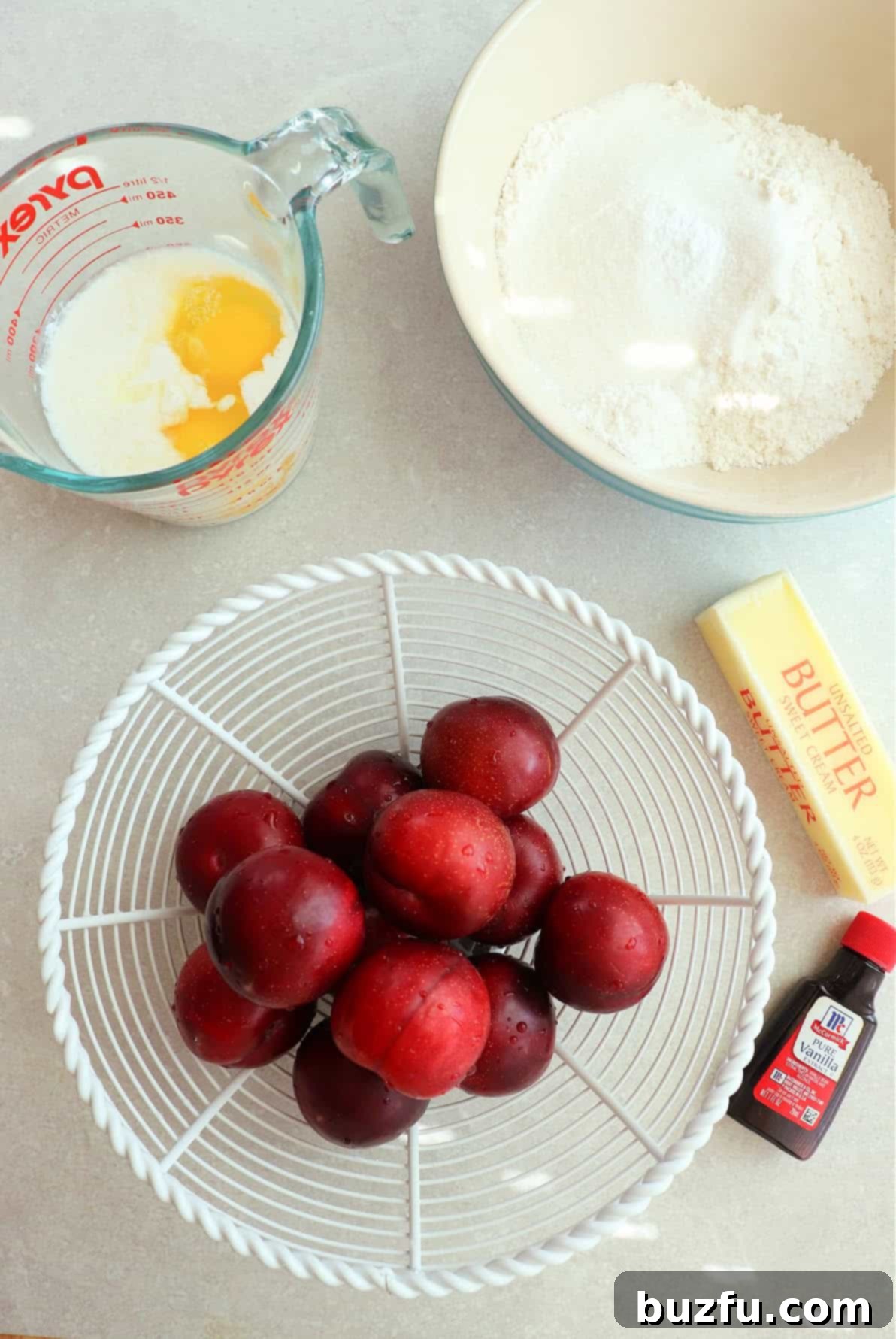 Dry cake ingredients in a bowl, a bottle of vanilla extract, stick of butter, buttermilk with eggs in a glass measuring cup and fresh plums in a white basket.