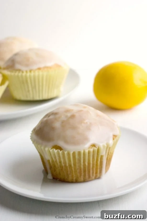 A beautifully glazed lemon muffin in a yellow cupcake wrapper, resting on a pristine white plate, showcasing its soft texture and inviting glaze.