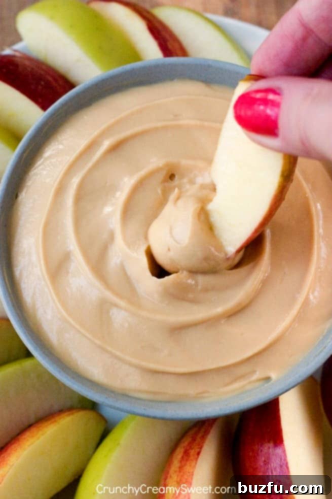 Creamy Caramel Apple Dip: An Addictive Two-Ingredient Treat Close-up of a hand dipping a slice of green apple into a rich, creamy caramel cheesecake dip, showcasing the perfect texture and inviting indulgence.