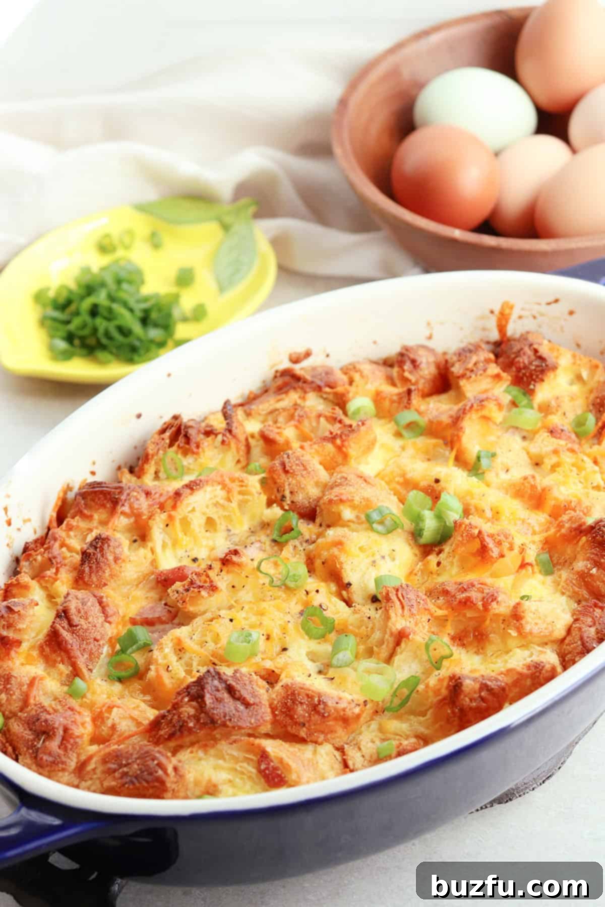 Bread and egg casserole in an oval baking dish with eggs in a wooden bowl behind it.