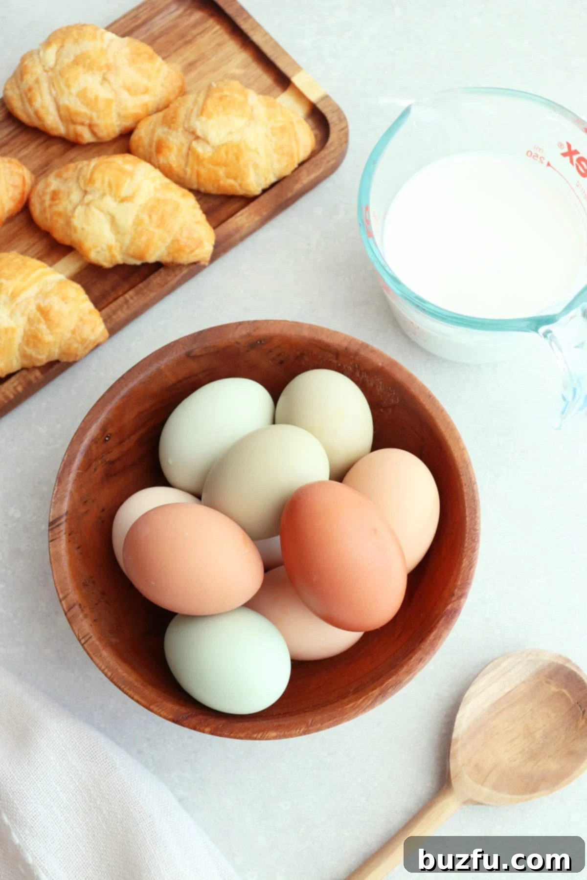 Eggs in a wooden bowl, mini croissants in a wooden tray, and milk in a glass measuring cup on a gray board.